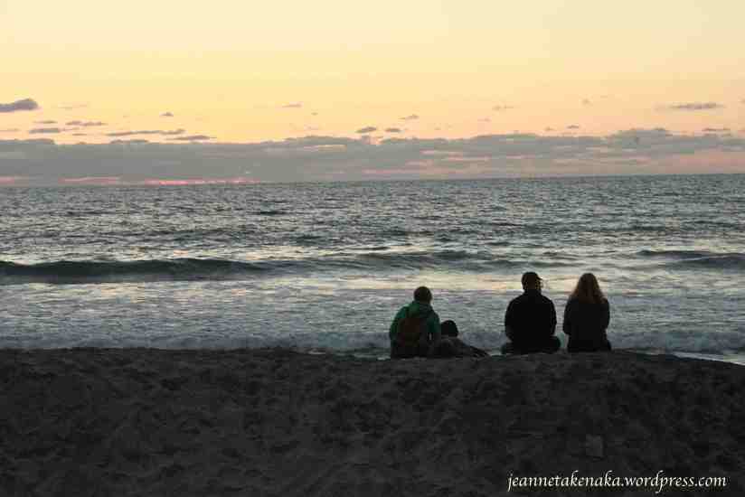 A man, woman and children sitting on the sand near the ocean at sunset