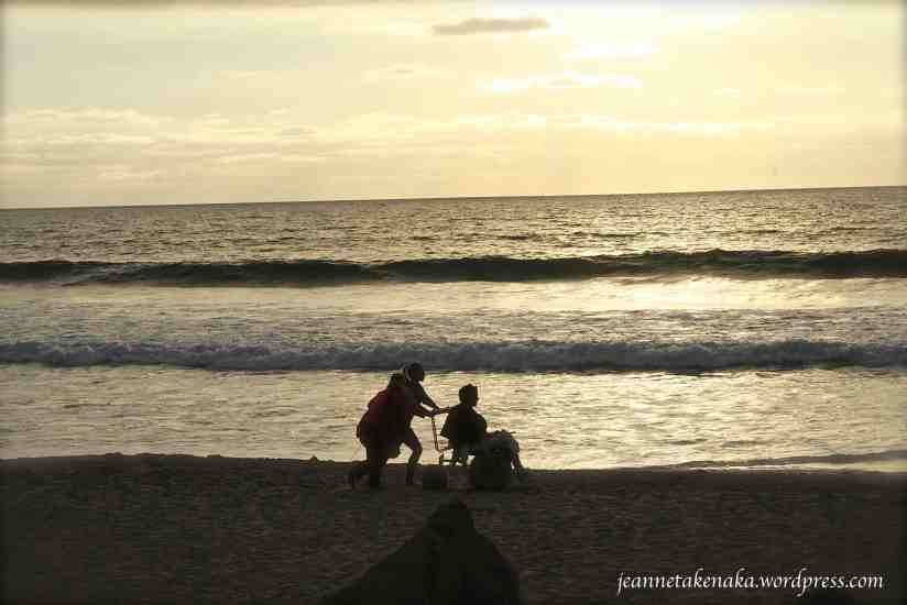 Two young women pushing a boy in a wheelchair on the beach at sunset