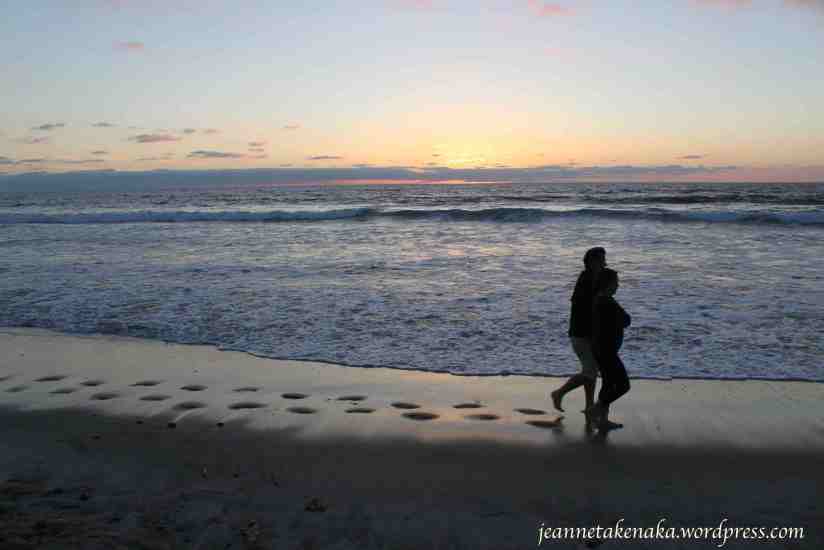 A husband and wife walking on the beach at sunset, their footprints trailing behind them on the wet sand